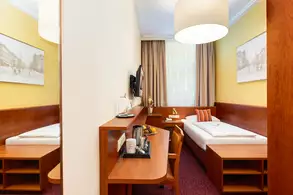 Ein einfaches Hotelzimmer mit einem Einzelbett, einem Schreibtisch und einem Fernseher an der Wand. Die Wände sind gelb gestrichen, und es gibt große Fenster mit Vorhängen. Auf dem Tisch stehen Obst und Getränke.