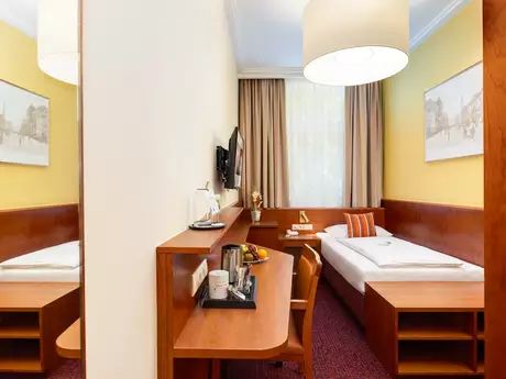 Ein einfaches Hotelzimmer mit einem Einzelbett, einem Schreibtisch und einem Fernseher an der Wand. Die Wände sind gelb gestrichen, und es gibt große Fenster mit Vorhängen. Auf dem Tisch stehen Obst und Getränke.