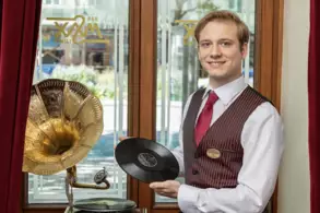 Ein junger Mann in Anzug steht neben einem Grammophon und hält eine Schallplatte in der Hand. Im Hintergrund sind große Fenster mit einem Blick auf die Außenansicht sowie rote Vorhänge zu sehen.
