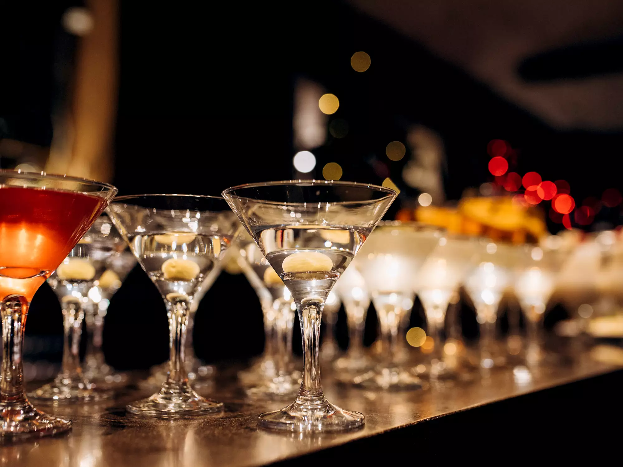 Eine Reihe von eleganten Cocktails in Gläsern steht auf einer Theke. Die Getränke variieren in Farbe und Art, mit einer warmen Beleuchtung im Hintergrund, die eine festliche Atmosphäre schafft.