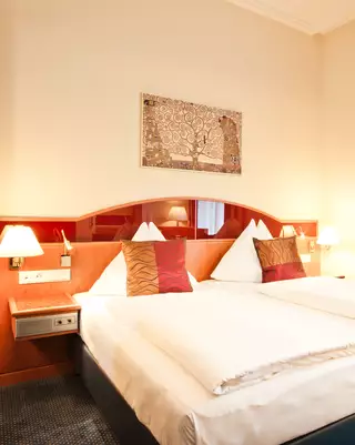 Ein helles Hotelzimmer mit zwei Einzelbetten, drei dekorativen Kopfkissen und Nachttischen. An der Wand hängt ein Bild. Die Fenster sind mit Vorhängen geschmückt. Eine Klimaanlage ist sichtbar, und das Zimmer hat einen Teppichboden.