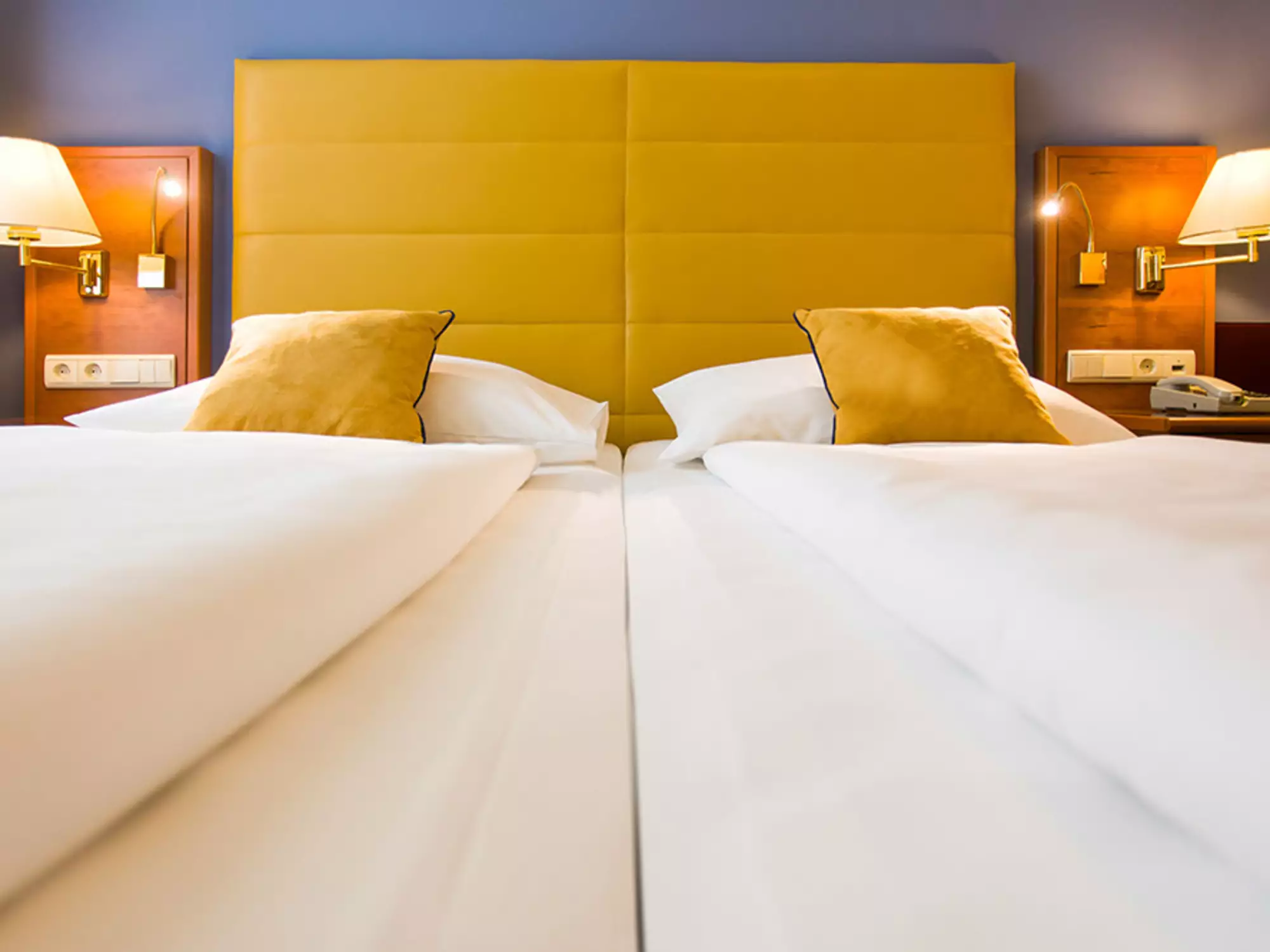 Das Bild zeigt ein Zimmer mit zwei Einzelbetten, die mit weißen Bettläufern und gelben Kissen dekoriert sind. Hinter den Betten befindet sich ein gelbes Kopfteil. An der Wand hängen zwei Nachttischlampen.