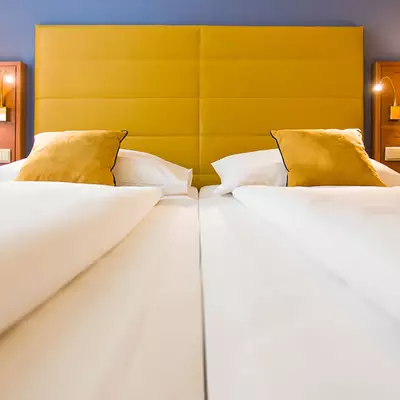 Das Bild zeigt ein Zimmer mit zwei Einzelbetten, die mit weißen Bettläufern und gelben Kissen dekoriert sind. Hinter den Betten befindet sich ein gelbes Kopfteil. An der Wand hängen zwei Nachttischlampen.