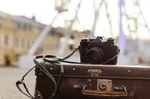 Eine analoge Kamera liegt auf einem alten Koffer, im Hintergrund sind unscharf ein Freizeitpark und ein Riesenrad zu erkennen.
