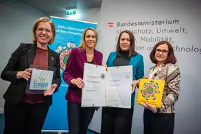 Vier Frauen stehen lächelnd vor einer Wand mit dem Schriftzug "Bundesministerium für Klimaschutz, Umwelt, Energie, Mobilität, Innovation und Technologie". Sie halten verschiedene Dokumente und ein Schild mit dem Schriftzug "Ecolabel". Im Hintergrund ist eine Informationsgrafik zu sehen.