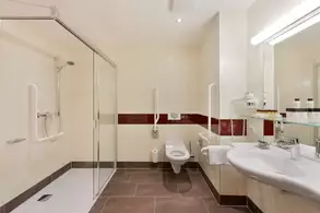 Sauberes Badezimmer mit einer Duschkabine, einem Waschbecken und einer Toilette. Es gibt ein großes Spiegel und Haltegriffe für zusätzlichen Halt. Die Wände sind hell beige mit einem schmalen roten Farbstreifen. Der Boden ist mit dunklen Fliesen ausgelegt.