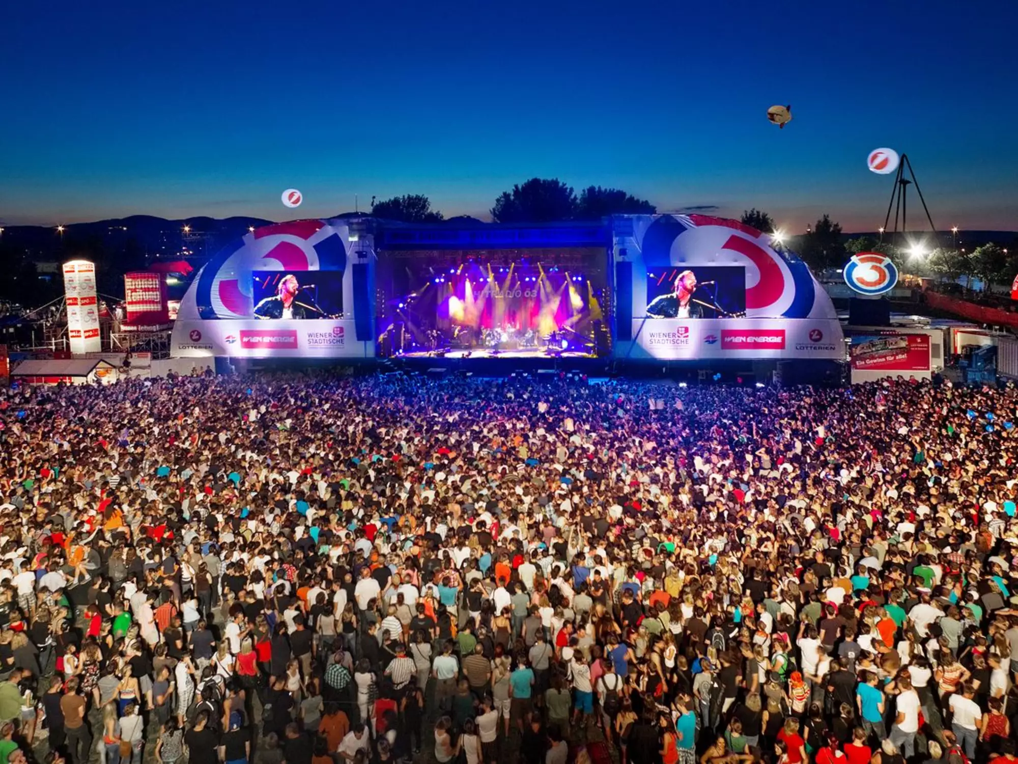 Eine große Menschenmenge versammelt sich vor einer Bühne bei einem Konzert oder Festival. Die Bühne ist beleuchtet und zeigt eine Performance, während der Himmel bei Sonnenuntergang leuchtet. Umgebende Strukturen und Veranstaltungselemente sind sichtbar.
