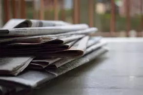 Ein Haufen gefalteter Zeitungen liegt auf einem Tisch. Die Zeitungen sind teilweise übereinander gestapelt, mit lesbaren Überschriften und Textausschnitten auf den obersten Seiten. Der Hintergrund ist unscharf, mit grünen und braunen Farbtönen.