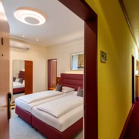Blick durch einen offenen Türrahmen in einen Hotelzimmerflur. Im Vordergrund ist ein Raum mit zwei Betten, einem Spiegel und einem Klimagerät sichtbar. Der Flur hat einen roten Teppich und gelbe Wände, mit Türen zu weiteren Zimmern auf beiden Seiten.