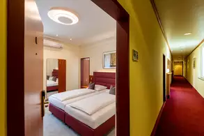 Blick durch einen offenen Türrahmen in einen Hotelzimmerflur. Im Vordergrund ist ein Raum mit zwei Betten, einem Spiegel und einem Klimagerät sichtbar. Der Flur hat einen roten Teppich und gelbe Wände, mit Türen zu weiteren Zimmern auf beiden Seiten.