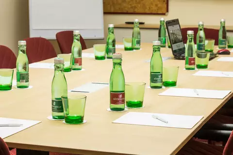 Ein Konferenztisch mit mehreren grünen Gläsern und Flaschen Wasser steht in einem Raum. Auf dem Tisch liegen Notizblätter und ein Laptop. Der Raum ist hell und modern gestaltet.
