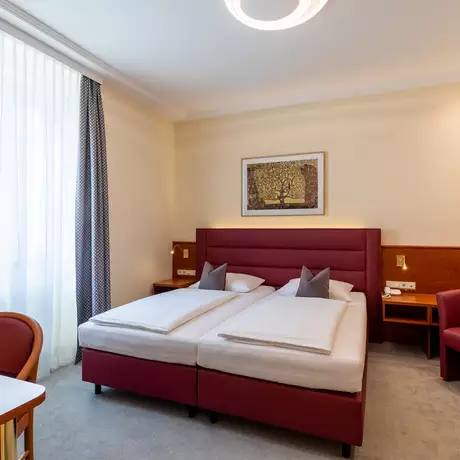 Ein gemütliches Hotelzimmer mit zwei Einzelbetten, einem roten Kopfteil und zwei Kissen. Im Raum gibt es einen Tisch aus Holz, einen roten Sessel und große Fenster mit Vorhängen. An der Wand hängt ein Bild mit natürlichen Motiven. Ambientebeleuchtung sorgt für eine angenehme Atmosphäre.