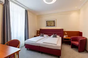 Ein gemütliches Hotelzimmer mit zwei Einzelbetten, einem roten Kopfteil und zwei Kissen. Im Raum gibt es einen Tisch aus Holz, einen roten Sessel und große Fenster mit Vorhängen. An der Wand hängt ein Bild mit natürlichen Motiven. Ambientebeleuchtung sorgt für eine angenehme Atmosphäre.