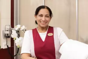 Eine lächelnde Frau in einem roten Kleid mit einem Namensschild steht in einem Badezimmer. Neben ihr sind weiße Orchideen und ein Haartrockner an der Wand zu sehen. Sie hält ein weißes Handtuch.