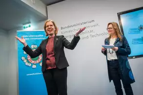 Eine Frau mit Brille und blonder Haarfarbe steht auf einer Bühne, lächelt und gestikuliert mit den Händen. Neben ihr steht eine weitere Frau, die ein Blatt Papier in der Hand hält. Im Hintergrund sind Symbole und Texte an der Wand zu sehen, die mit Umweltschutz in Verbindung stehen.