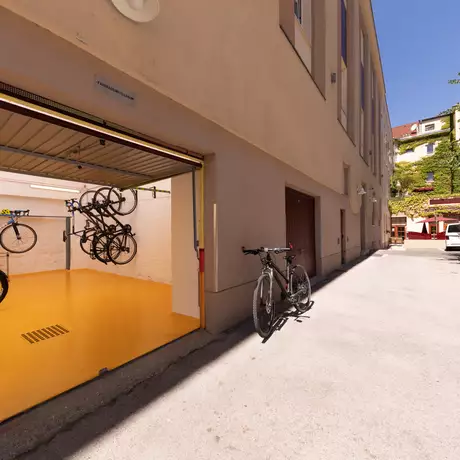Ein Fahrradabstellraum mit mehreren Fahrrädern an Wänden aufgehängt. Der Boden ist in einem leuchtenden Gelb gestrichen. Rechts davon steht ein weiteres Fahrrad auf dem Bürgersteig. Im Hintergrund ist ein Teil eines mehrstöckigen Gebäudes und einige Autos zu sehen.