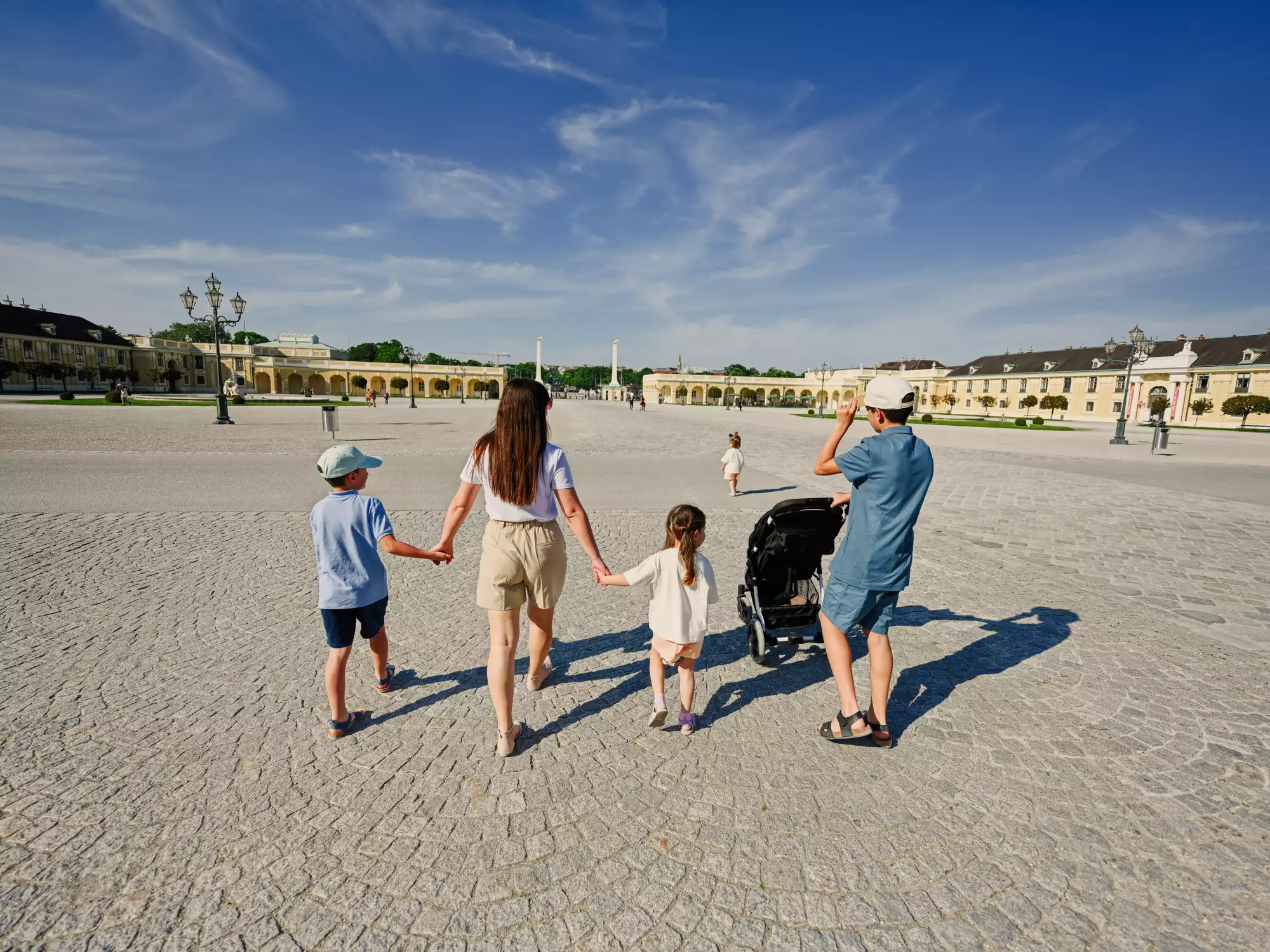 Eine Familie spaziert über einen weitläufigen Platz. Die Mutter hält die Hände zweier Kinder, während ein weiteres Kind im Kinderwagen sitzt. Im Hintergrund sind historische Gebäude und ein blauer Himmel sichtbar.