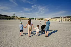 Eine Familie spaziert über einen weitläufigen Platz. Die Mutter hält die Hände zweier Kinder, während ein weiteres Kind im Kinderwagen sitzt. Im Hintergrund sind historische Gebäude und ein blauer Himmel sichtbar.