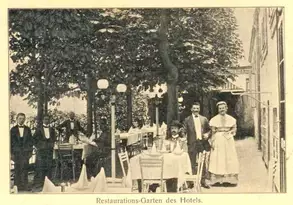 Ein Restaurantgarten mit mehreren Tischen und Stühlen im Freien. Im Vordergrund stehen mehrere Kellner in formeller Kleidung und eine Frau in einem Kleid. Bäume und Laternen sind im Hintergrund sichtbar, was eine angenehme Atmosphäre schafft.