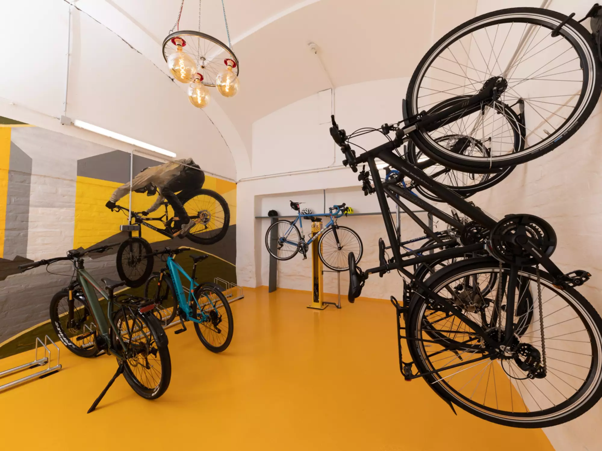 Ein Bicycle-Showroom mit mehreren Fahrrädern, die an der Wand hangen und auf Ständern stehen. Die Wände sind gelb und mit einem grauen Bergmotiv dekoriert. Ein eleganter Kronleuchter hängt von der Decke.