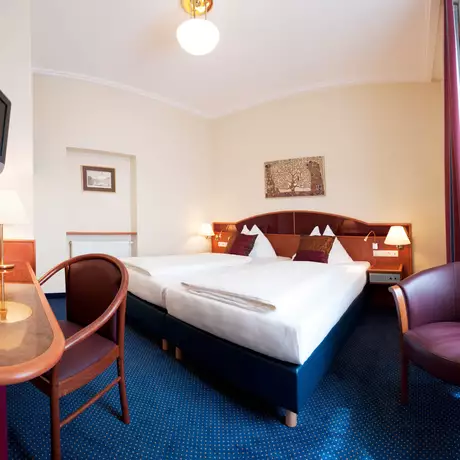 Ein Hotelzimmer mit zwei Betten, einer kleinen Lampe auf einem Tisch und einem Fernseher an der Wand. Zwei Stühle stehen in der Ecke des Raumes, der mit Teppichboden ausgelegt ist. Es gibt einen Fenster mit Vorhängen und einige dekorative Elemente an der Wand.