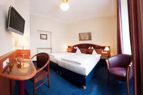 Ein Hotelzimmer mit zwei Betten, einer kleinen Lampe auf einem Tisch und einem Fernseher an der Wand. Zwei Stühle stehen in der Ecke des Raumes, der mit Teppichboden ausgelegt ist. Es gibt einen Fenster mit Vorhängen und einige dekorative Elemente an der Wand.