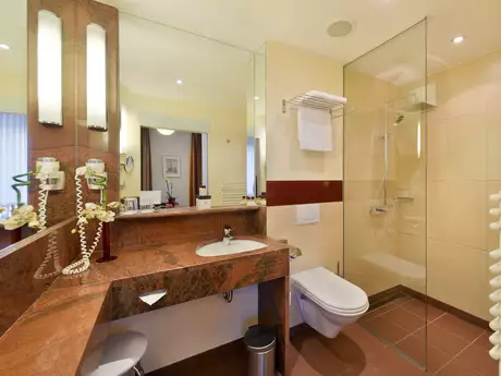 Ein modernes Badezimmer mit einem Waschtisch aus Marmor, Spiegel und einer Dusche aus Glas. Es gibt einen Toilettenbereich, Handtücher hängen an der Wand, und neben dem Waschbecken stehen Pflegeprodukte in einem dekorativen Arrangement. Sanfte Beleuchtung schafft eine angenehme Atmosphäre.
