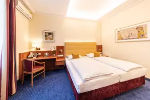 Ein modernes Hotelzimmer mit einem Doppelbett, einer gemütlichen Sitzgelegenheit und einem Schreibtisch. An der Wand sind Kunstwerke und eine Lampe angebracht. Der Boden ist mit blauen Teppichfliesen bedeckt, und große Fenster lassen viel Licht herein.