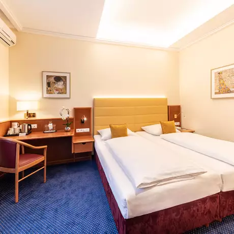 Ein modernes Hotelzimmer mit einem Doppelbett, einer gemütlichen Sitzgelegenheit und einem Schreibtisch. An der Wand sind Kunstwerke und eine Lampe angebracht. Der Boden ist mit blauen Teppichfliesen bedeckt, und große Fenster lassen viel Licht herein.
