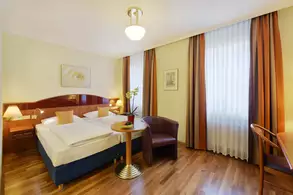 Das Bild zeigt ein modernes Hotelzimmer mit zwei Betten, einem Tisch mit Obst und einer Pflanze, sowie einem Sessel. Die Wände sind hell gehalten und die Vorhänge haben orangefarbene Streifen. Es gibt eine Wandlampe und ein kleines Kunstwerk. Der Boden ist aus Holz.