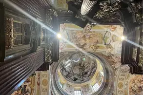 Blick in die Decke einer prunkvollen Kirche mit kunstvoller Malerei und Verzierungen. Die runde Kuppel ist sichtbar, umgeben von detaillierten Wandfresken und goldenen Elementen. Helle Lichter betonen die kunstvollen Details im Innenraum.