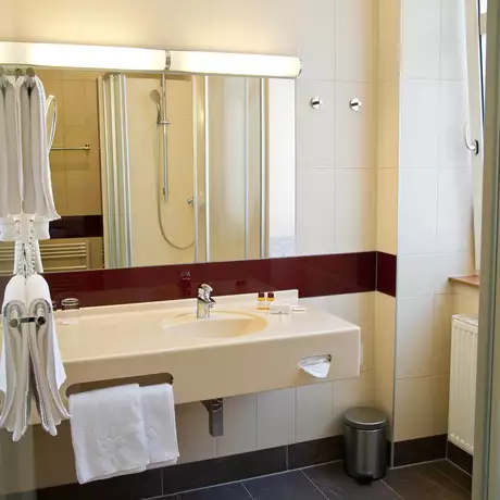 Ein modernes Badezimmer mit einem Waschbecken, einem Spiegel und einer Dusche. An der Wand hängen Handtücher, und auf der Ablage befinden sich Pflegeprodukte. Der Boden ist gefliest, und es gibt einen kleinen Müllbehälter.