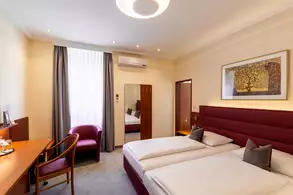 Ein modernes Hotelzimmer mit zwei Einzelbetten, einem Schreibtisch, einem Stuhl und einem roten Sessel. Große Fenster mit Vorhängen lassen Tageslicht herein. An der Wand hängt ein Rahmendes Bild, und ein Spiegel steht gegenüber der Betten.