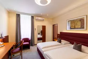 Ein modernes Hotelzimmer mit zwei Einzelbetten, einem Schreibtisch, einem Stuhl und einem roten Sessel. Große Fenster mit Vorhängen lassen Tageslicht herein. An der Wand hängt ein Rahmendes Bild, und ein Spiegel steht gegenüber der Betten.
