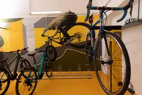 In einem modernen Fahrradladen stehen mehrere Fahrräder auf einem orangefarbenen Boden. An der Wand befindet sich ein großes Wandgemälde eines Radfahrers in Aktion. Im Hintergrund sind eine Fahrradpumpe und ein Fahrradständer sichtbar.