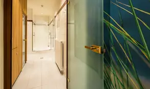 Derbild zeigt einen modernen Badezimmerflur mit einer gläsernen Tür, die zu einer Dusche führt. Die Wände sind hell gefliest, und es gibt einen Heizkörper. Im Hintergrund ist eine Wand mit einem Motiv von Gräsern zu sehen.