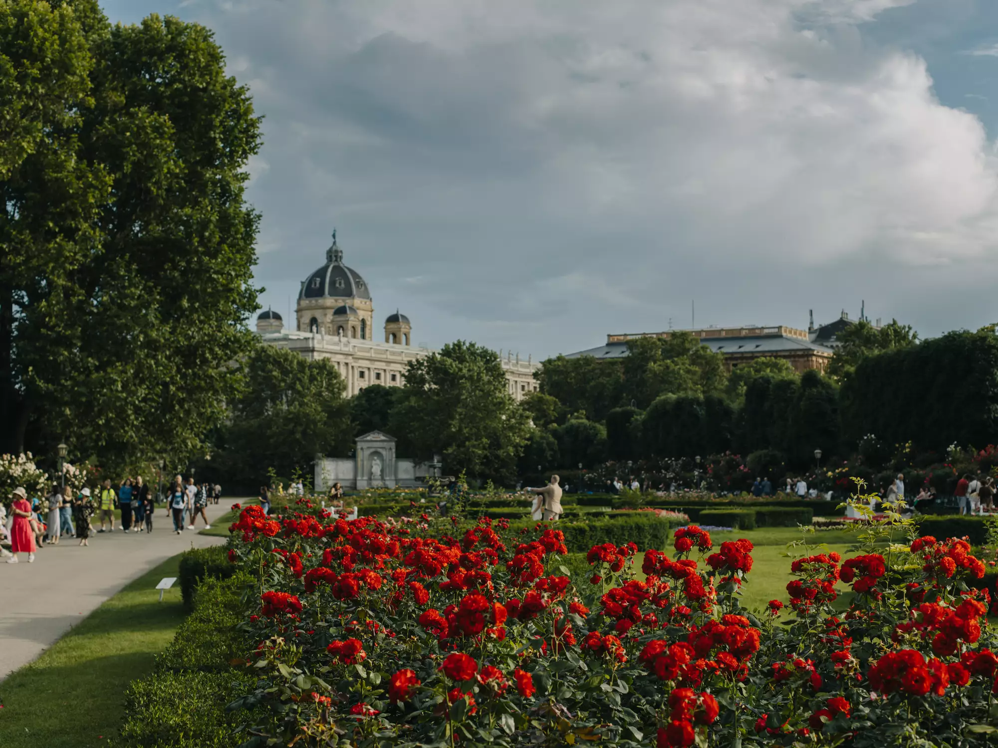 Blick auf einen Garten mit roten Blumen im Vordergrund und grünen Bäumen im Hintergrund. Im Hintergrund sind Menschen zu sehen, die im Park spazieren gehen, sowie ein historisches Gebäude mit einer Kuppel. Der Himmel ist bewölkt.