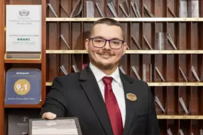 Ein Hotelmitarbeiter steht an der Rezeption und lächelt freundlich. Er trägt einen Anzug mit einer roten Krawatte und hält eine Mappe in der Hand. Im Hintergrund sind mehrere Fächer mit Informationen und Auszeichnungen des Hotels sichtbar.
