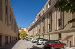 Ein schmaler Innenhof mit hohen, modernen Gebäuden auf beiden Seiten. Parkende Autos säumen den Weg. Im Hintergrund sind bunte Fenster und Pflanzen sichtbar, der Himmel ist blau und sonnig.