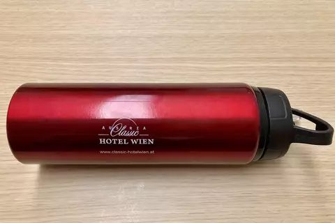 Ein roter Trinkbecher mit schwarzem Deckel, auf dem "Austria Classic Hotel Wien" und die Website gedruckt sind. Der Becher liegt auf einer hellen Holzoberfläche.