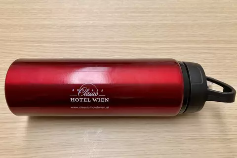 Ein roter Trinkbecher mit schwarzem Deckel, auf dem "Austria Classic Hotel Wien" und die Website gedruckt sind. Der Becher liegt auf einer hellen Holzoberfläche.