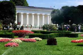 Ein gepflegter Park mit bunten Blumenbeeten und grünen Rasenflächen. Im Hintergrund steht ein klassisches Gebäude mit Säulen, umgeben von Besuchern. Es sind auch Bäume und Bewässerungssysteme sichtbar.