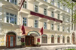 Die Außenansicht eines Hotels mit dem Schriftzug "Hotel Wien" über dem Eingang. Die Fassade ist hell und hat große Fenster. Rote Fahnen hängen an der Front. Der Eingangsbereich ist mit Pflanzen dekoriert.