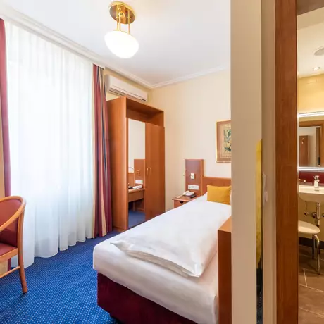 Ein Hotelzimmer mit einem Einzelbett, einem Schreibtisch, einem Stuhl und einer Fußmatte. Im Hintergrund sind ein angrenzendes Badezimmer mit Dusche und ein großes Fenster mit Vorhängen zu sehen. Die Wände sind in warmen Farben gehalten und das Zimmer ist gut beleuchtet.