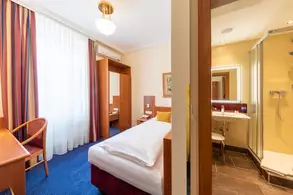 Ein Hotelzimmer mit einem Einzelbett, einem Schreibtisch, einem Stuhl und einer Fußmatte. Im Hintergrund sind ein angrenzendes Badezimmer mit Dusche und ein großes Fenster mit Vorhängen zu sehen. Die Wände sind in warmen Farben gehalten und das Zimmer ist gut beleuchtet.