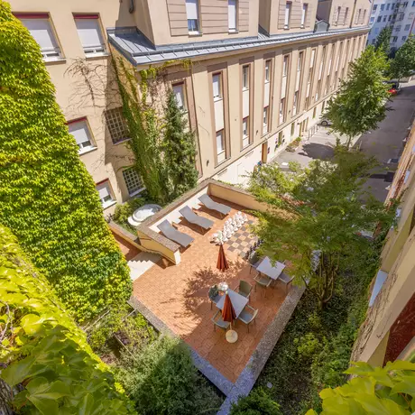 Blick auf einen ruhigen Innenhof eines Gebäudes, umgeben von hohen Wänden und begrünten Pflanzen. Im Innenhof sind einige Liegen, Sonnenschirme und Sitze auf einem gepflasterten Bereich angeordnet, umgeben von grüner Vegetation.