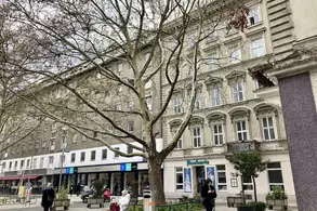 Stadtplatz mit einem großen Baum im Vordergrund. Im Hintergrund sind mehrere Gebäude mit Fensterfronten sichtbar, darunter ein Geschäft. Einige Passanten gehen vorbei, und auf einer Bank sitzt eine Person im Rollstuhl. Die Atmosphäre wirkt urban und geschäftig.