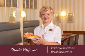 Eine lächelnde Mitarbeiterin des Frühstücksservice trägt einen Teller mit frischem Obst. Im Hintergrund sind Tische und Stühle eines Restaurantbereichs sichtbar, mit warmen Farben und einer gemütlichen Atmosphäre.