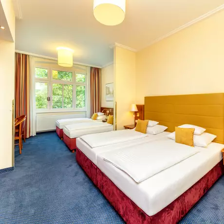 Ein helles Hotelzimmer mit zwei Einzelbetten, einem kleinen Tisch und einem Stuhl. Große Fenster lassen viel Licht herein. Der Boden ist mit Teppich ausgelegt und es gibt einen Kleiderschrank sowie Nachttische mit Lampen.