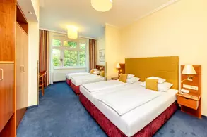 Ein helles Hotelzimmer mit zwei Einzelbetten, einem kleinen Tisch und einem Stuhl. Große Fenster lassen viel Licht herein. Der Boden ist mit Teppich ausgelegt und es gibt einen Kleiderschrank sowie Nachttische mit Lampen.